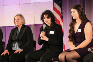 one school board member, seated left, beside two student panelists with the one in the middle holding a microphone