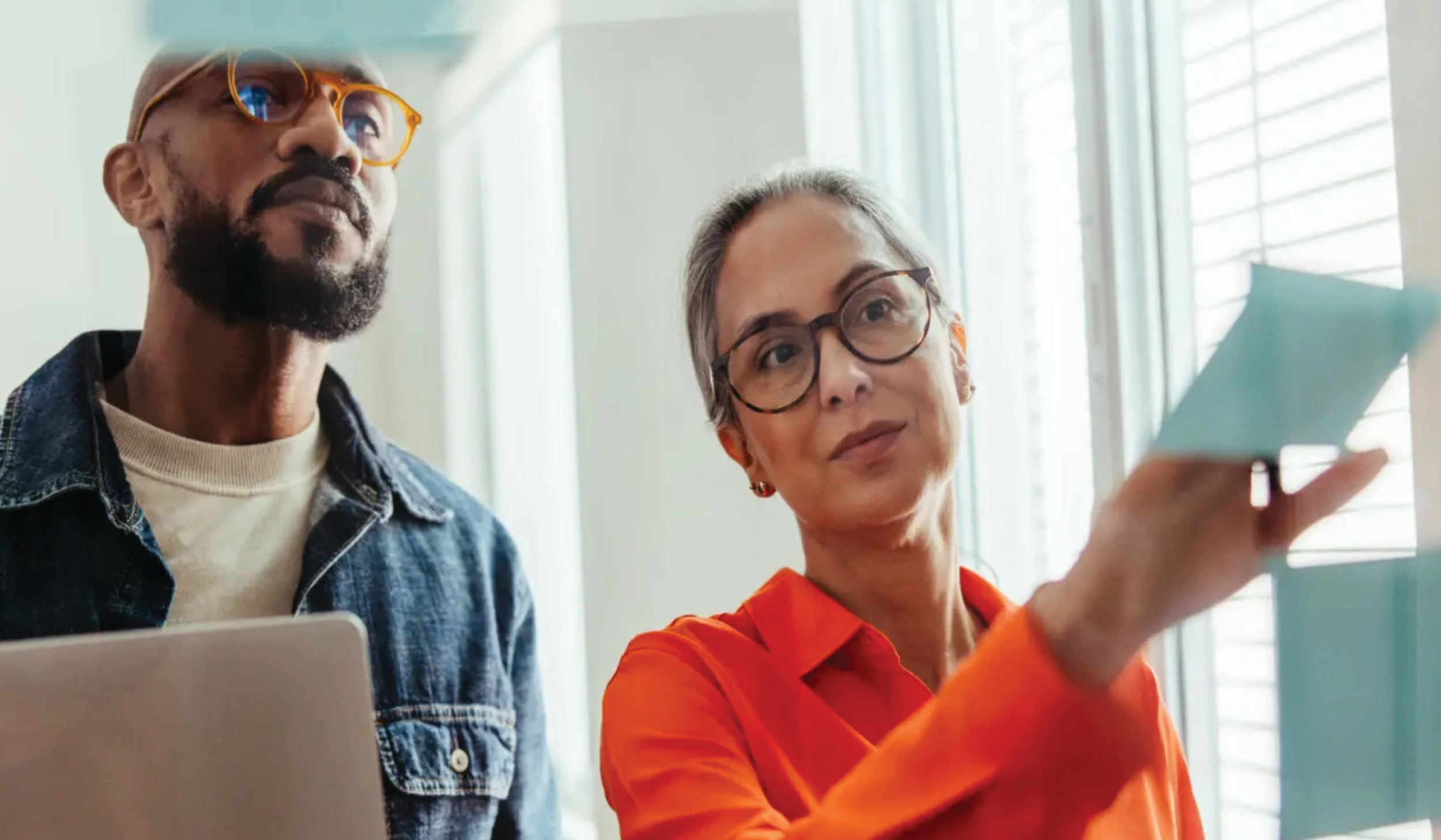 Two people looking at and placing sticky notes on a transparent glass wall.