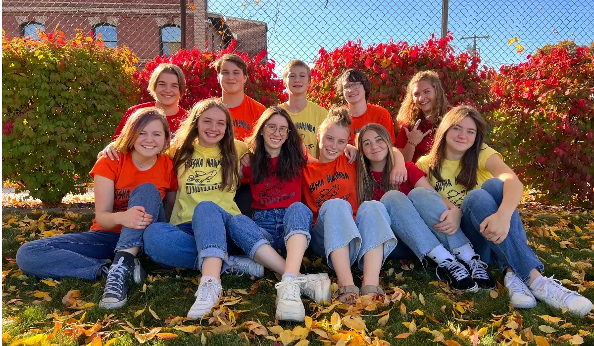 Members of the Musha Marimba student ensemble seated together outdoors in autumn, wearing group t-shirts